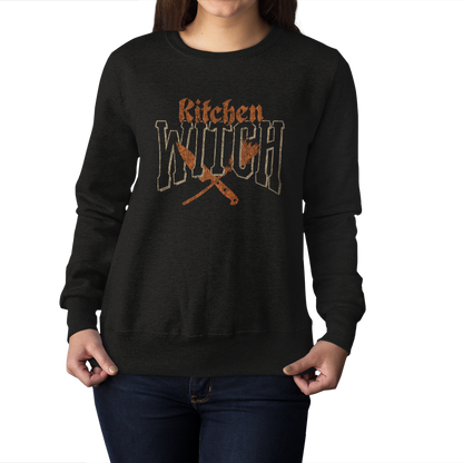 A woman wearing a black crewneck sweatshirt with a distressed orange graphic that reads 'Kitchen Witch' above crossed kitchen utensils, shown against a plain white background.