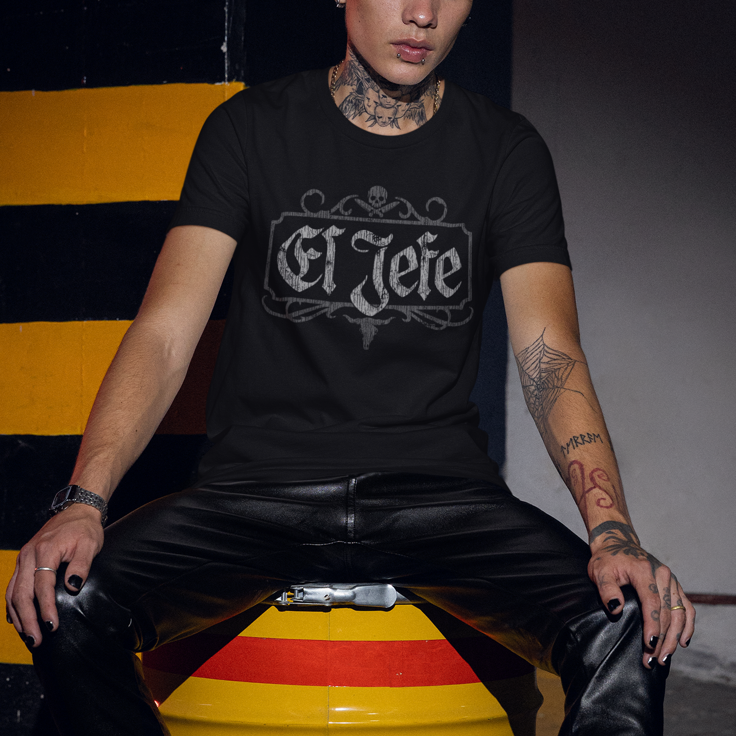 A tattooed man wearing a black graphic t-shirt with 'El Jefe' printed in a gothic font, black leather pants, and black boots, sitting on a yellow and red metal barrel in a dimly lit industrial setting.