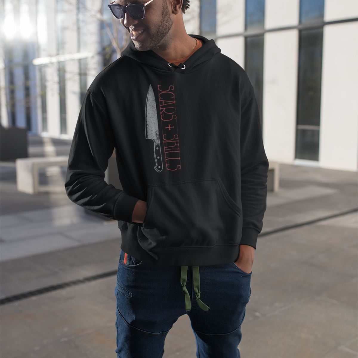 A man wearing a black hoodie with a graphic print of a knife and the text 'STALK + SHOOT' in red, paired with dark blue jeans and sunglasses, standing outdoors in front of a modern glass building.