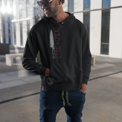A man wearing a black hoodie with a graphic print of a knife and the text 'STALK + SHOOT' in red, paired with dark blue jeans and sunglasses, standing outdoors in front of a modern glass building.