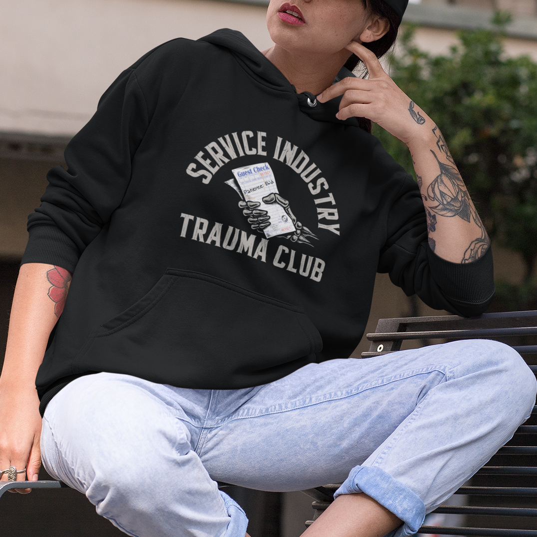 A person wearing a black hoodie with 'SERVICE INDUSTRY TRAUMA CLUB' printed on the front, light blue jeans, and a black cap, sitting on a bench in an outdoor urban setting.
