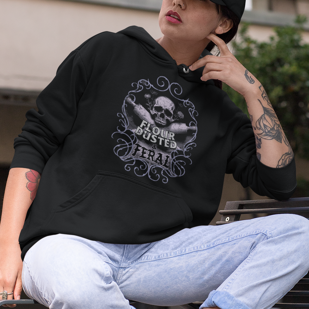 A person wearing a black graphic hoodie, light blue jeans, and a black cap sits on a metal bench in an urban outdoor setting with graffiti-covered buildings in the background.