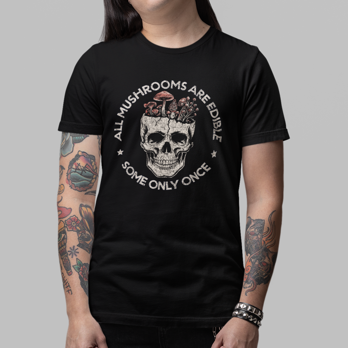 A model wearing a black graphic t-shirt with a skull and crossbones design and the text 'ALL MUSHROOMS ARE FATAL SOME ONLY ONCH', paired with black pants and boots, against a solid gray background.