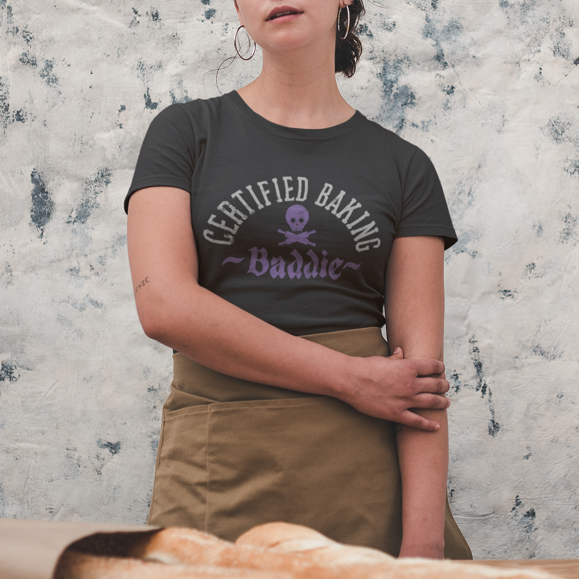 Person wearing a 'Certified Baking Buddy' t-shirt standing behind a table with bread, against a textured wall.
