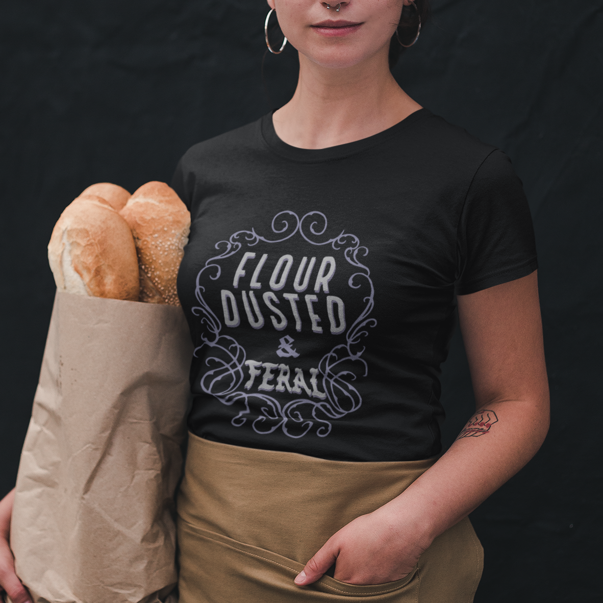 Person wearing a black t-shirt with 'Flour Dusted & Tearable' text, holding bread and a paper bag against a dark background.
