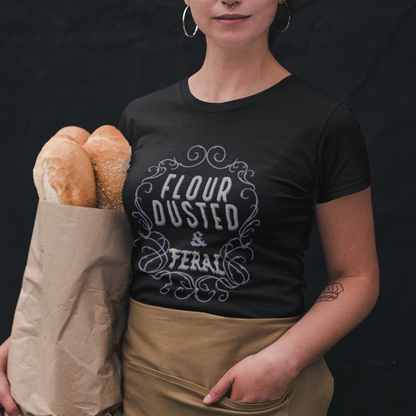 Person wearing a black t-shirt with 'Flour Dusted & Tearable' text, holding bread and a paper bag against a dark background.