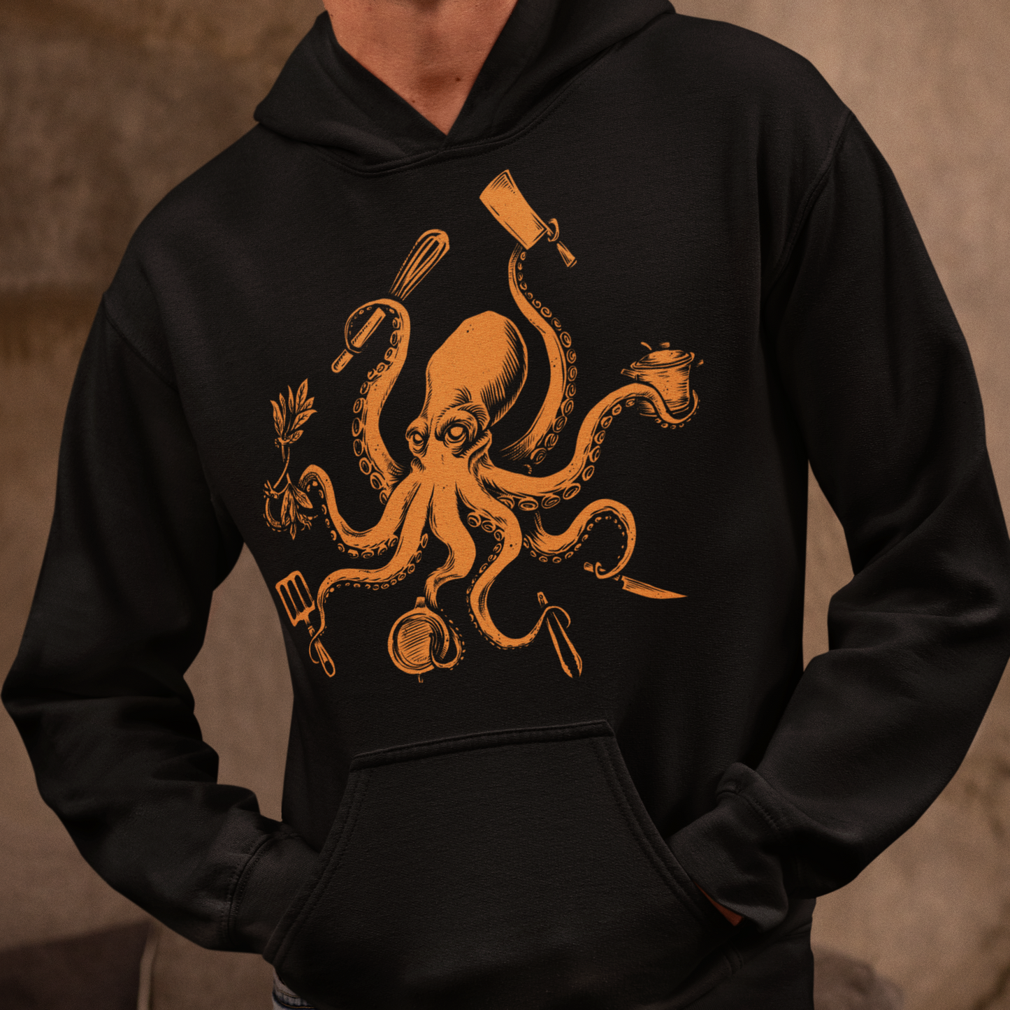 A man wearing a black pullover hoodie with a large orange graphic of an octopus surrounded by cooking utensils, paired with light blue jeans, set against a warm, textured brown background.