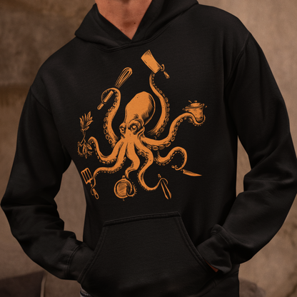 A man wearing a black pullover hoodie with a large orange graphic of an octopus surrounded by cooking utensils, paired with light blue jeans, set against a warm, textured brown background.