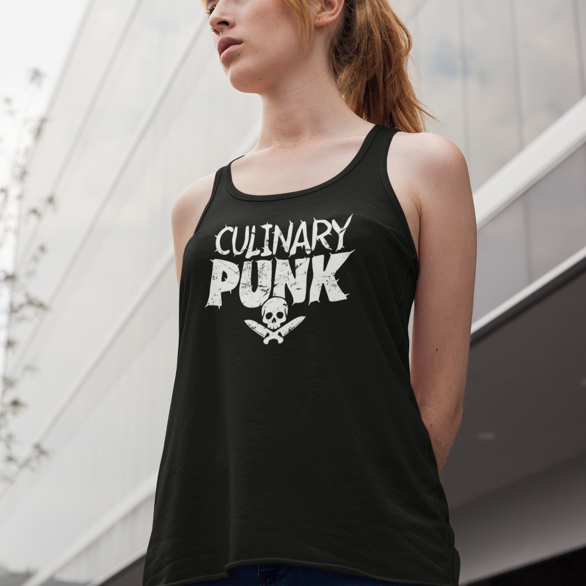 A woman wearing a black tank top with a white graphic that reads 'CULINARY PUNK' and a skull-and-crossbones design, photographed outdoors against a modern building background.