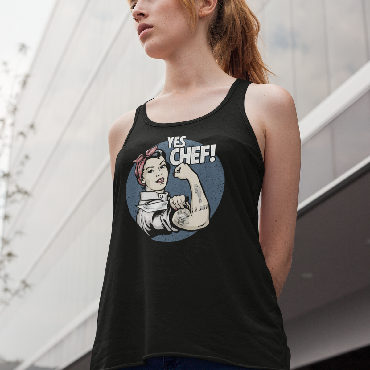 A woman wearing a black racerback tank top with a 'Yes Chef!' graphic inspired by Rosie the Riveter, photographed outdoors against a modern building facade.