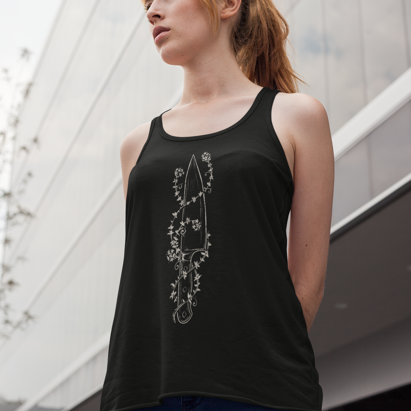A woman wearing a black tank top with a white floral dagger graphic, photographed outdoors against a modern glass building.