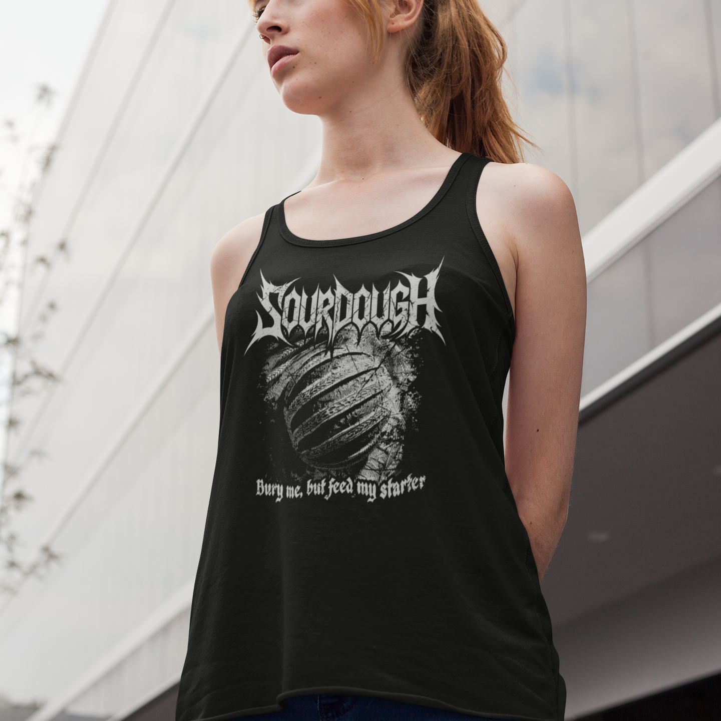 A woman wearing a black tank top with a white graphic print of the band Sourdough and the text 'Burn me, heat me, feed my hunger' in an outdoor urban setting.