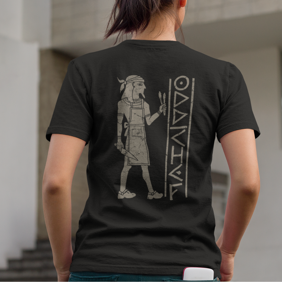 A woman viewed from behind wearing a black t-shirt with a large gray graphic of an ancient Egyptian figure and the word 'MAURITIUS' printed on the back, standing outdoors in front of a modern building.