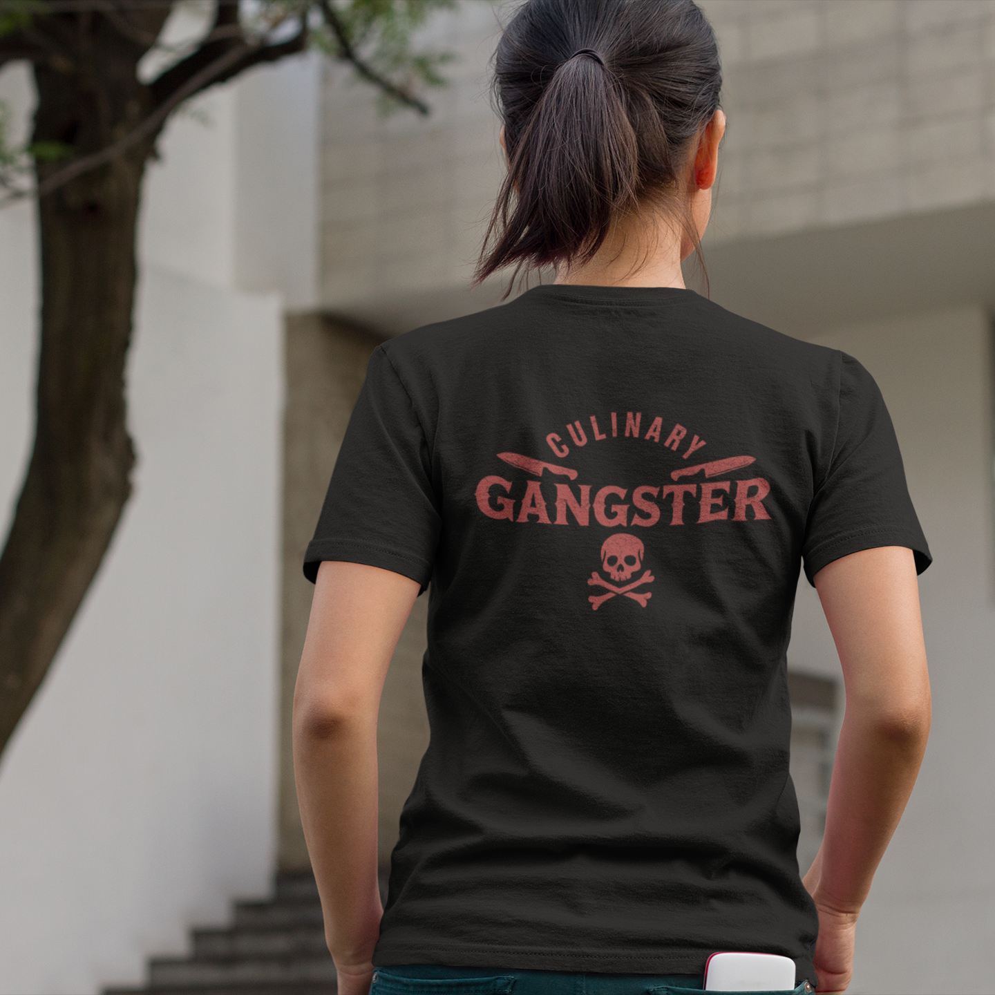 A woman viewed from behind wearing a black t-shirt with a red graphic that reads 'CULINARY GANGSTER' and a skull and crossbones, standing outdoors in front of a modern building.