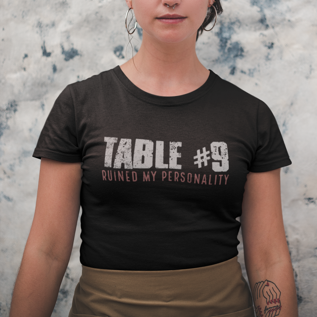 Person wearing a black t-shirt with 'TABLE #9 RUINED MY PERSONALITY' text against a textured wall.