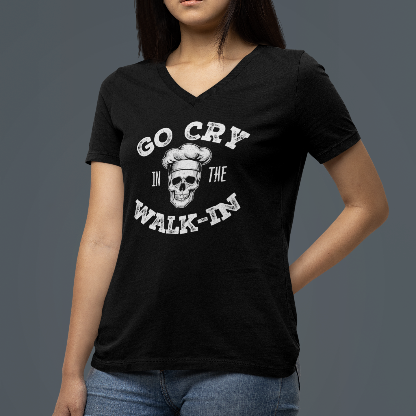 A woman wearing a black V-neck t-shirt with a white graphic print that reads 'GO CRY IN THE WALK-IN' around a skull wearing a chef's hat, paired with blue jeans, set against a solid dark gray background.