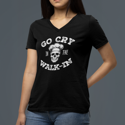 A woman wearing a black V-neck t-shirt with a white graphic print that reads 'GO CRY IN THE WALK-IN' around a skull wearing a chef's hat, paired with blue jeans, set against a solid dark gray background.