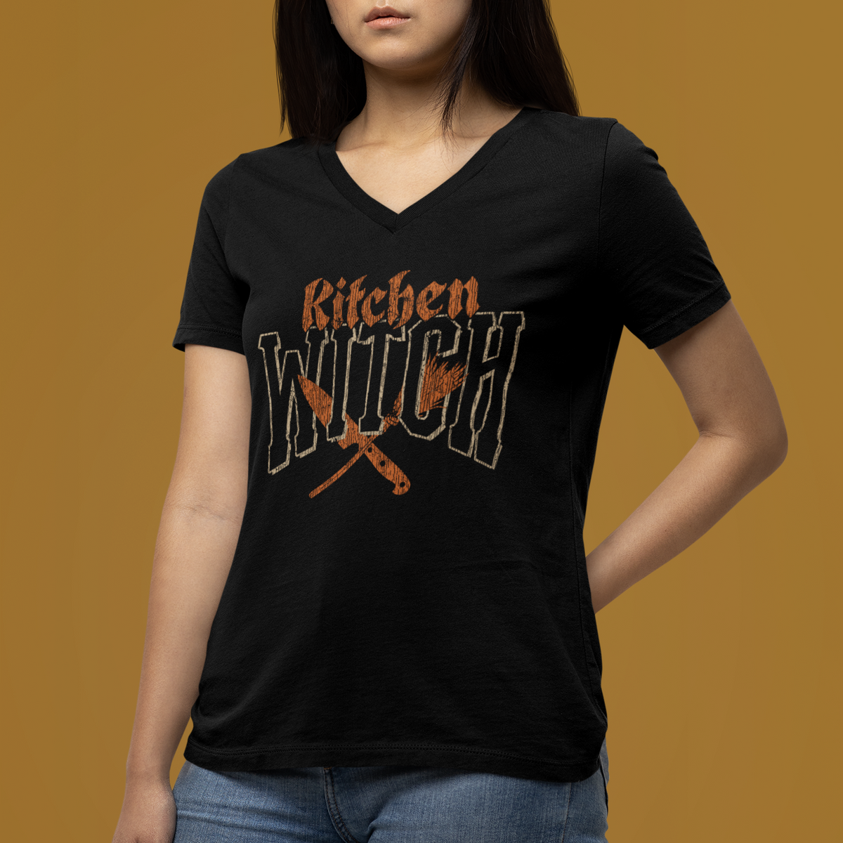 A woman wearing a black V-neck t-shirt with a distressed orange and white graphic that reads 'Kitchen Witch' with crossed kitchen tools, posed against a solid mustard yellow background.