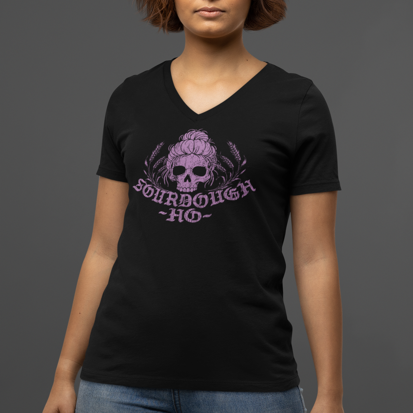 A woman wearing a black V-neck t-shirt with a purple skull graphic and the text 'SCHRBOUGHA HO', paired with blue jeans, against a solid dark gray background.