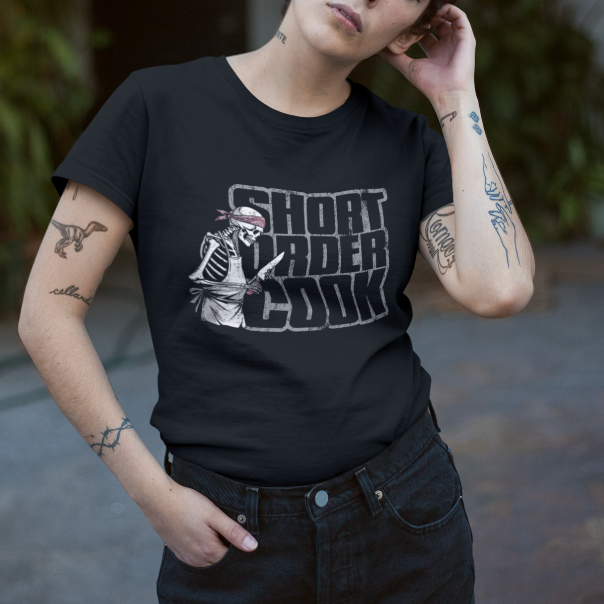 A person wearing a black graphic T-shirt with the text 'GHOST RANGER COOK' and a skeleton soldier illustration, paired with black denim shorts, standing in a softly blurred outdoor doorway with plants in the background.