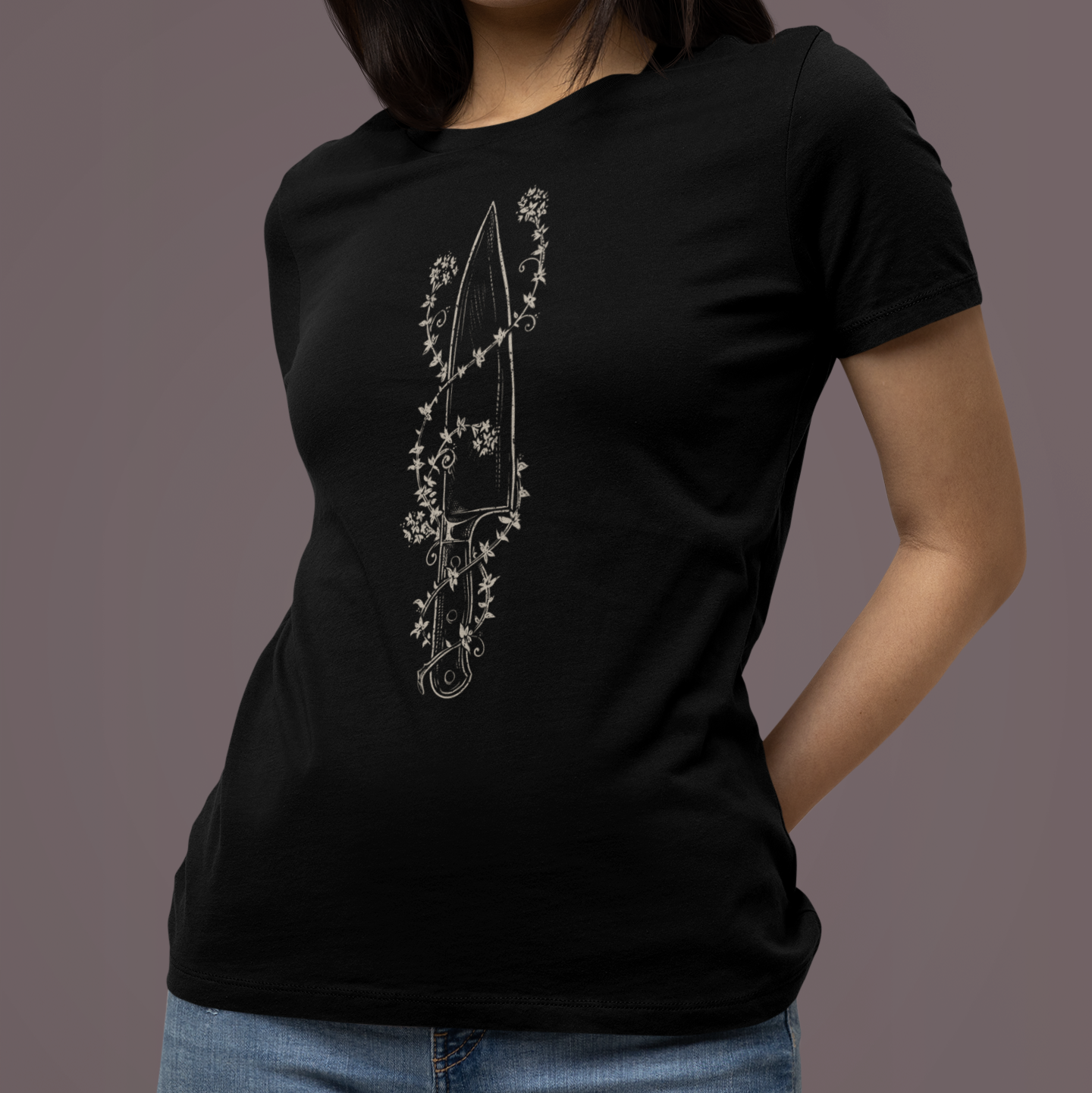 A woman wearing a black short-sleeve crewneck t-shirt with a white line-art graphic of a dagger surrounded by flowers on the front, paired with blue jeans, set against a solid dark gray studio background.