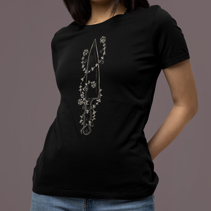 A woman wearing a black short-sleeve crewneck t-shirt with a white line-art graphic of a dagger surrounded by flowers on the front, paired with blue jeans, set against a solid dark gray studio background.