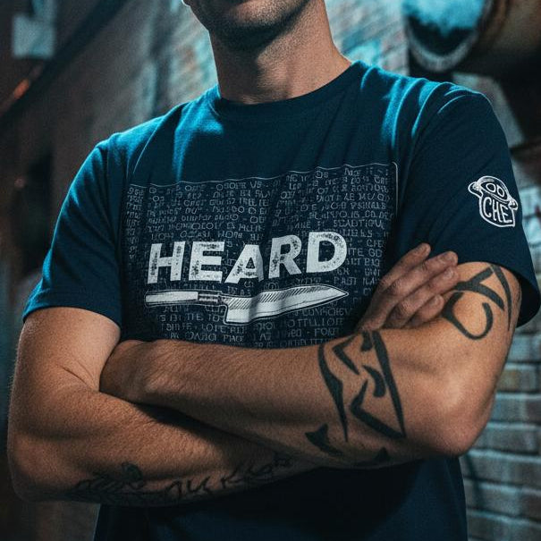 Man wearing a blue t-shirt with 'HEARD' text, standing against a graffiti-covered wall at night.