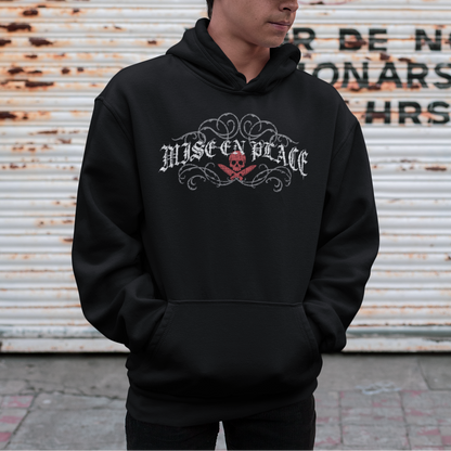 A man wearing a black hoodie with a 'Misan Peace' graphic on the chest, paired with a black baseball cap, standing in front of a rusty metal shutter with graffiti.