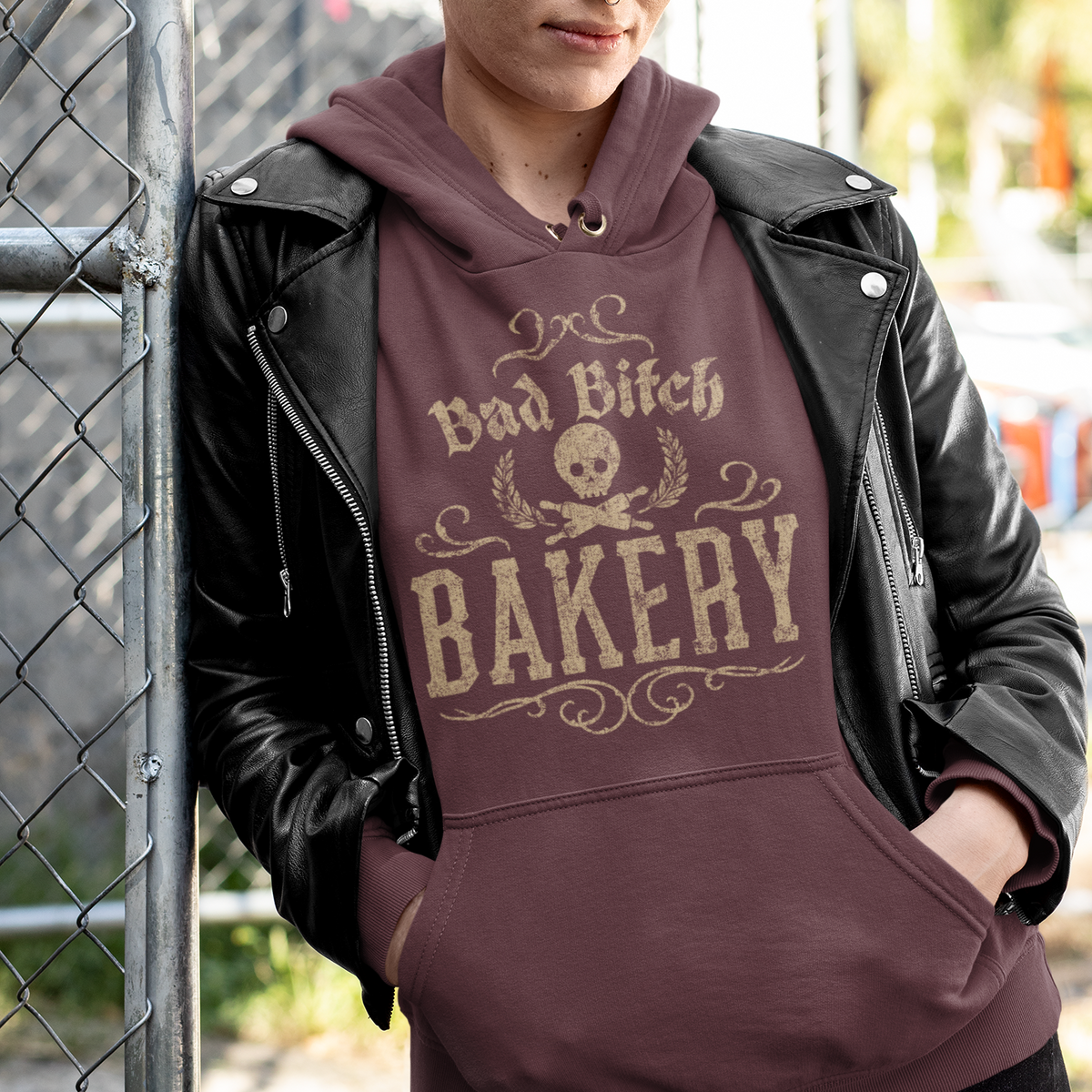 A person wearing a brown hoodie with a beige graphic that reads 'Bad Bitch BAKERY' and a skull design, layered under a black leather jacket, leaning against a chain-link fence outdoors.