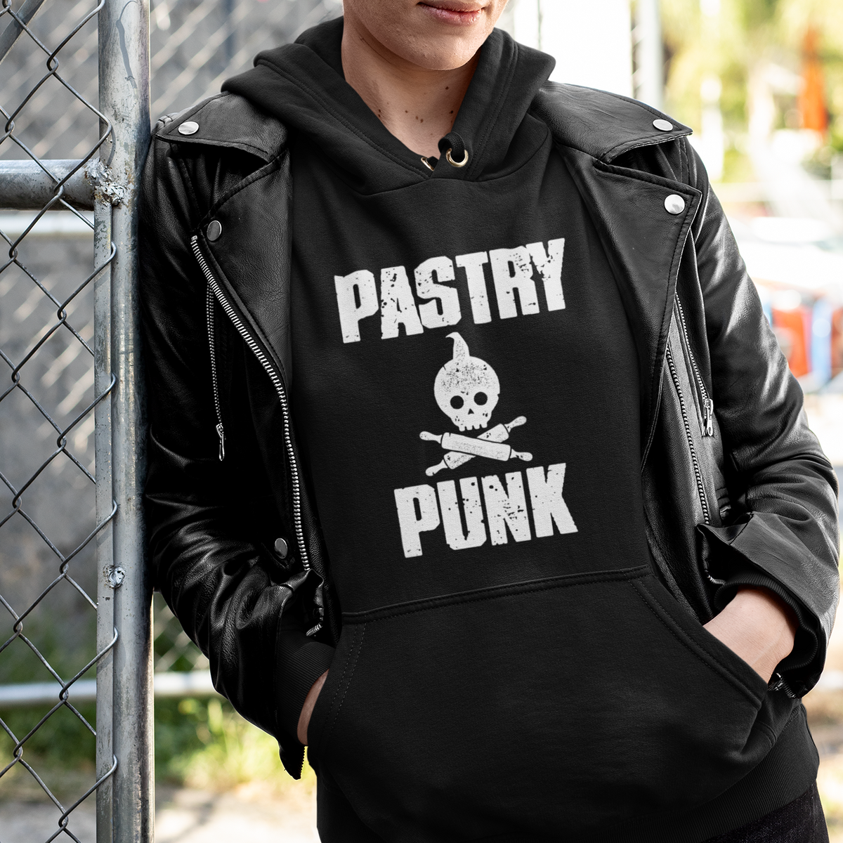 A person wearing a black hoodie with a white 'PASTRY PUNK' skull graphic, styled with a black leather jacket, leaning against a chain-link fence in an outdoor urban setting.