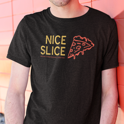 A smiling man wearing a black graphic t-shirt with 'NICE SLICE' and a pizza slice design, paired with bright red pants, standing against a pink tiled wall.