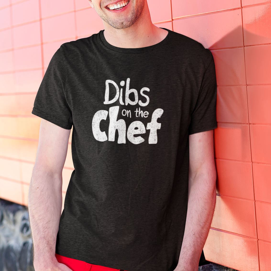 Man wearing a black t-shirt with 'Dibs on the Chef' text against a pink tiled wall.