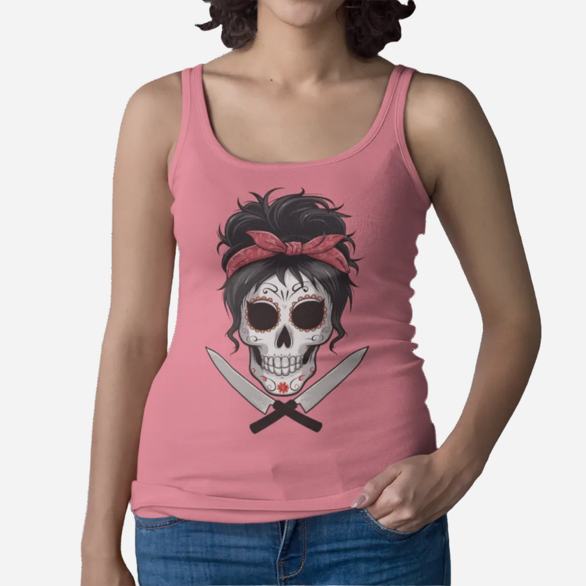 A woman wearing a pink sleeveless tank top with a graphic of a skull wearing a red bandana and crossed knives, paired with blue jeans against a plain white background.