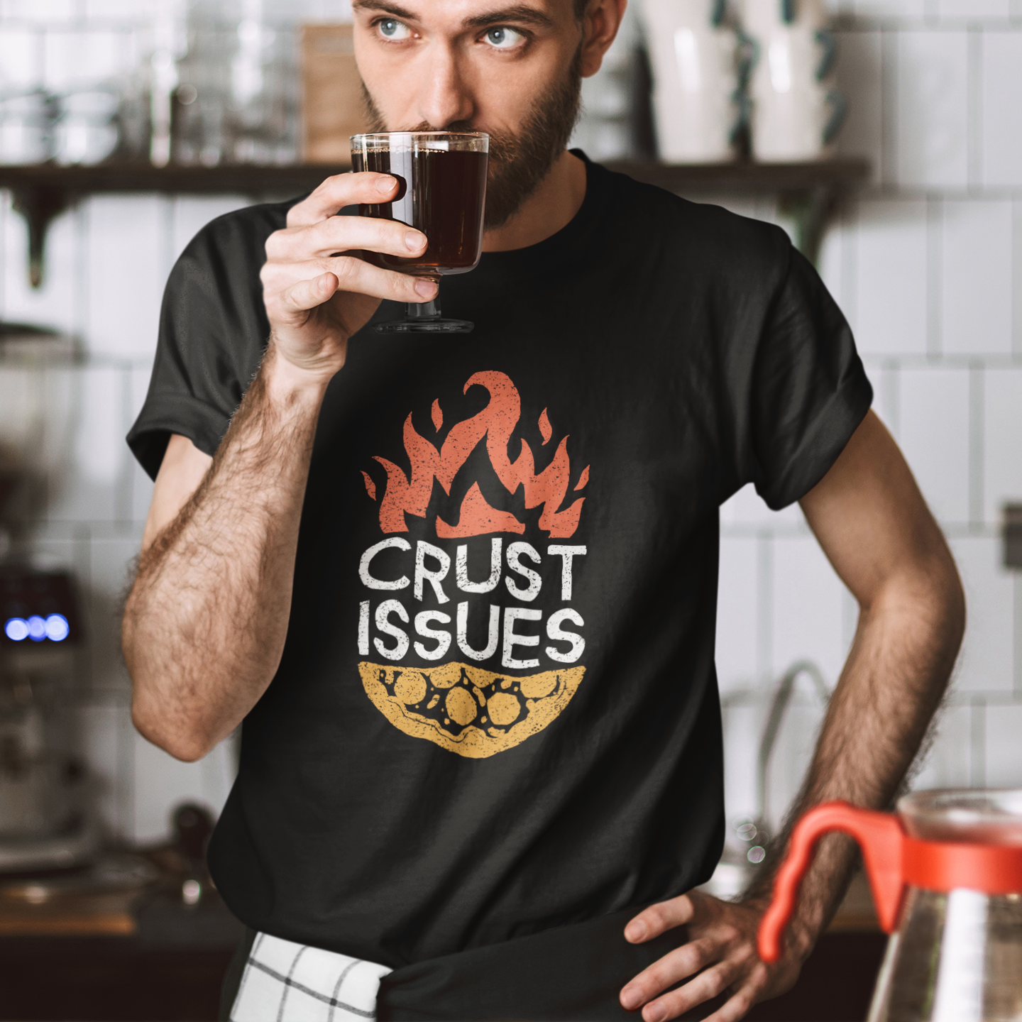 A man in a kitchen setting wearing a black graphic t-shirt with a flame and cookie design that reads 'CRUST ISSUES'. He is holding a glass of coffee.