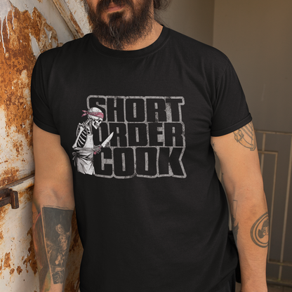 A man wearing a black graphic t-shirt with the text 'SHORT ORDER COOK' and a skeleton chef illustration, standing next to a rusted metal door.