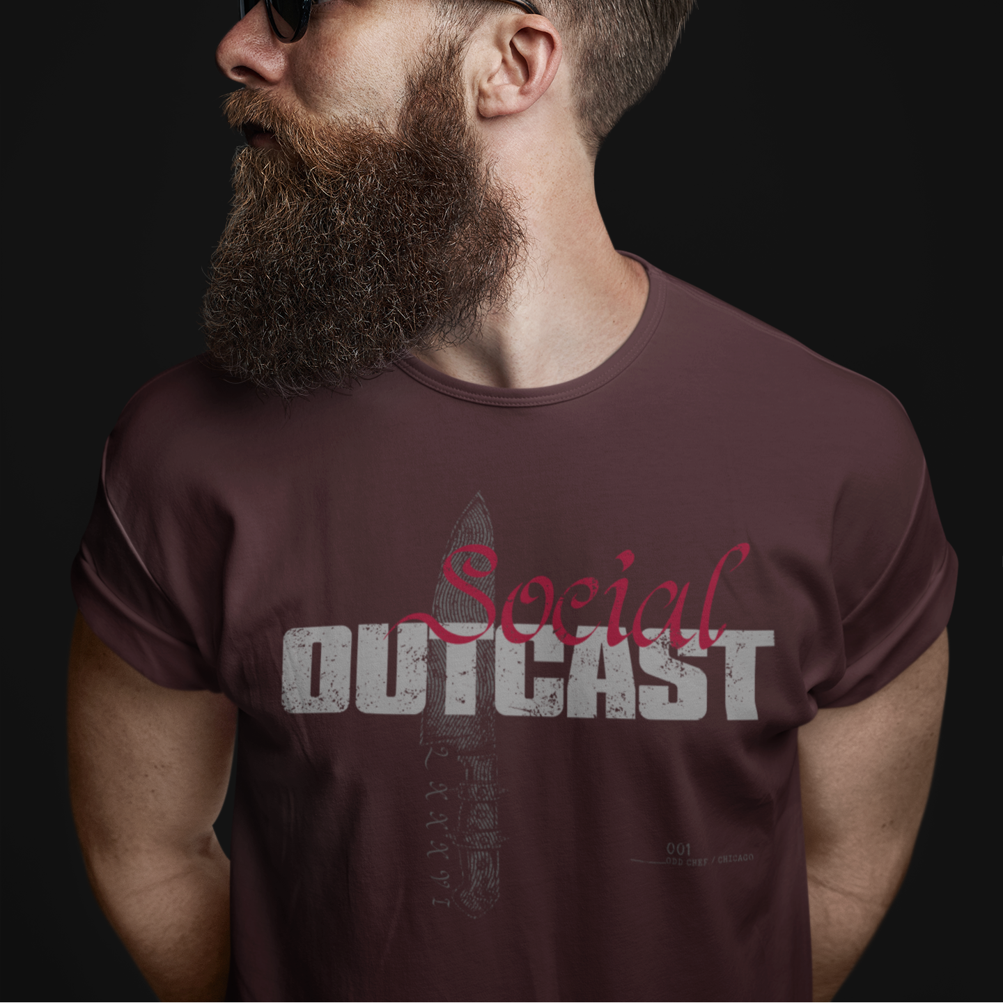 A man with a full beard and sunglasses wearing a dark brown t-shirt with a graphic print that reads 'Social OUTCAST' in distressed white and red lettering, set against a solid black background.