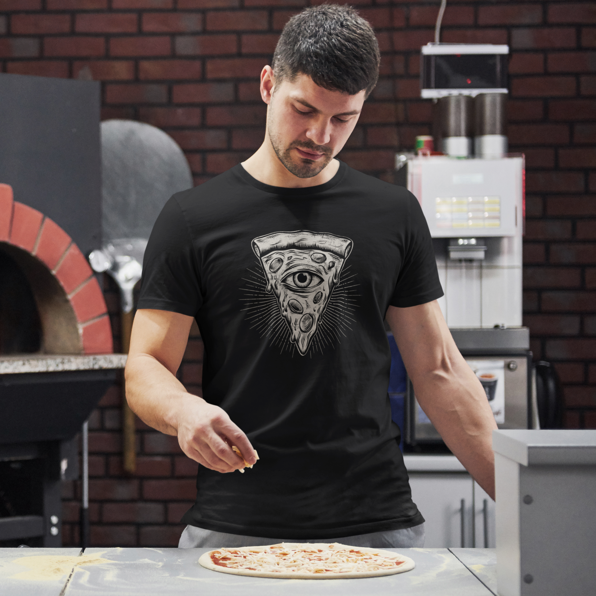 A man wearing a black t-shirt with a detailed grayscale pizza slice graphic featuring an all-seeing eye, standing in a kitchen and sprinkling cheese onto a pizza on a counter.