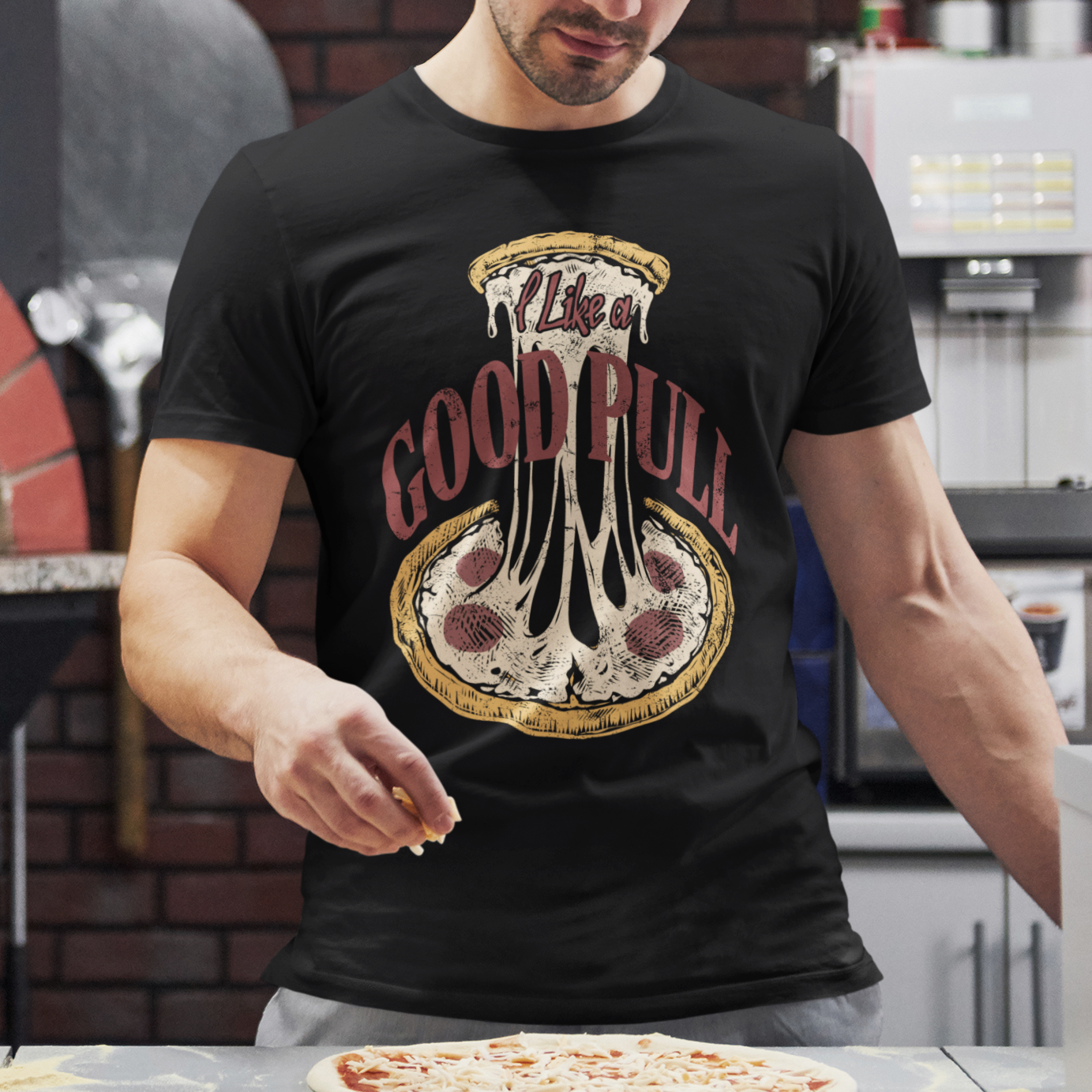A man wearing a black graphic t-shirt with a dripping pizza design and the text 'I live in GOOD RULL' stands in a pizza kitchen, sprinkling cheese onto a pizza on a counter.