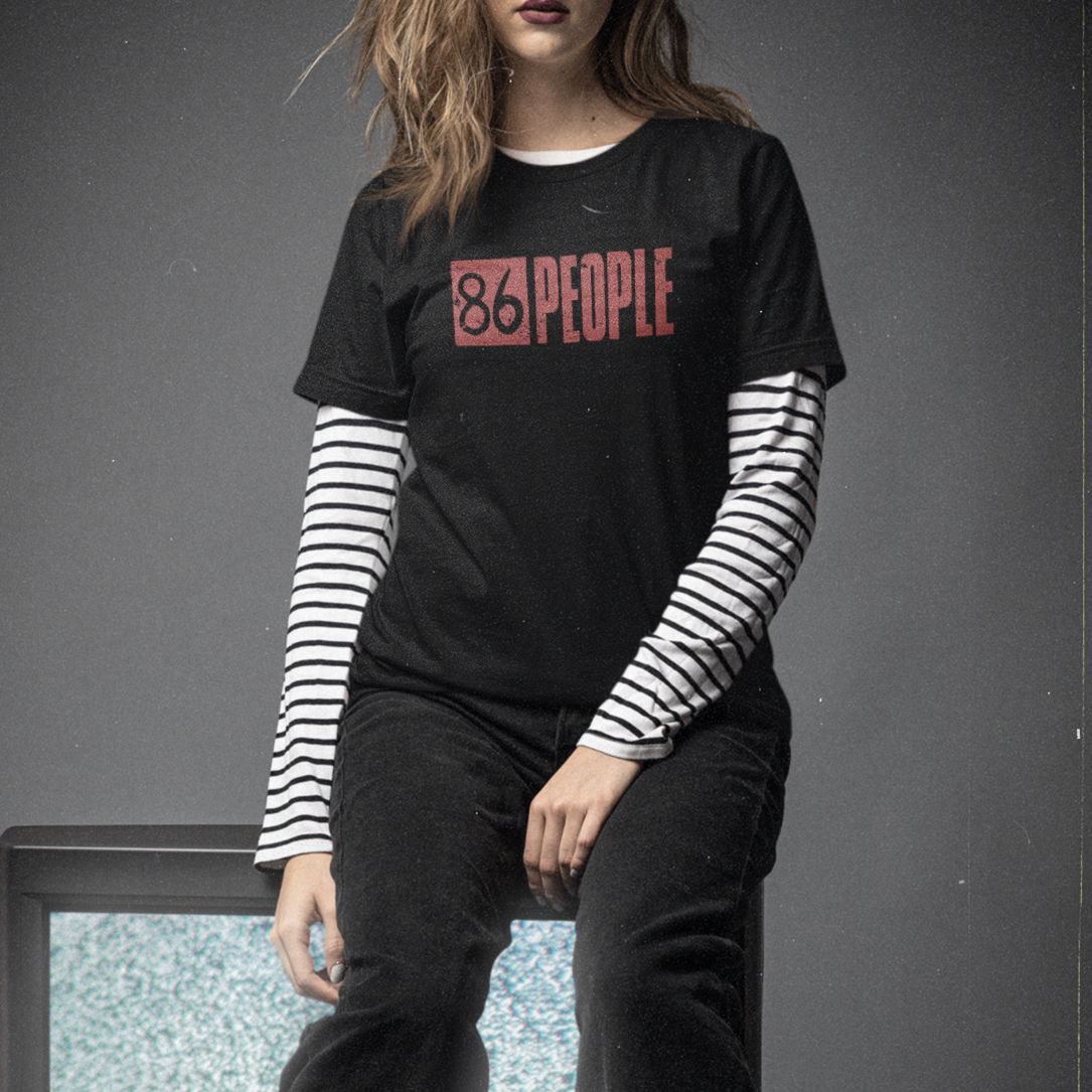 A woman wearing a black graphic t-shirt with '86 PEOPLE' printed in red, layered over a black and white striped long-sleeve shirt, dark jeans, and black boots, posing in front of a vintage television set against a dark gray studio background.