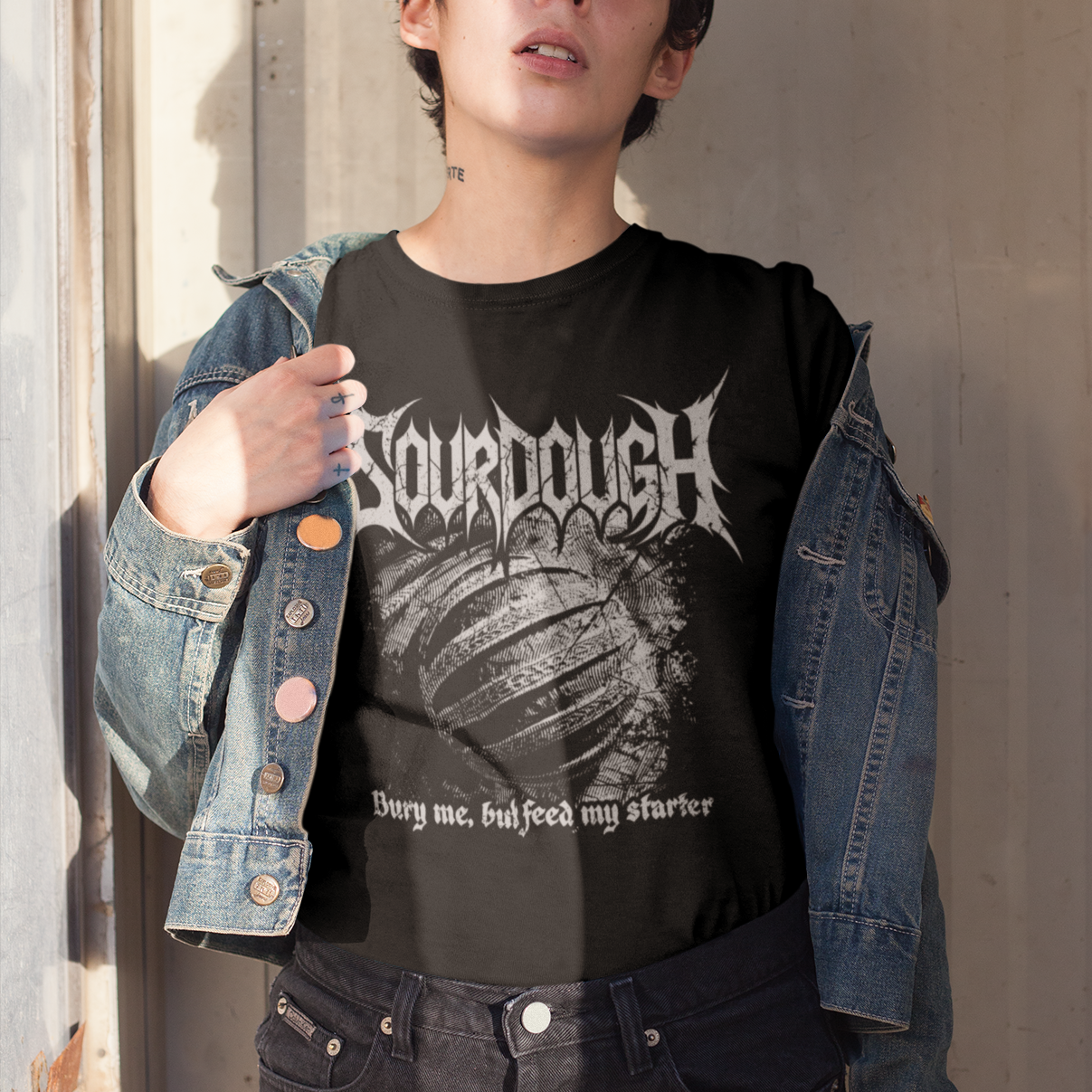 Person wearing a black t-shirt with 'Sourdough' design and denim jacket against a neutral wall.