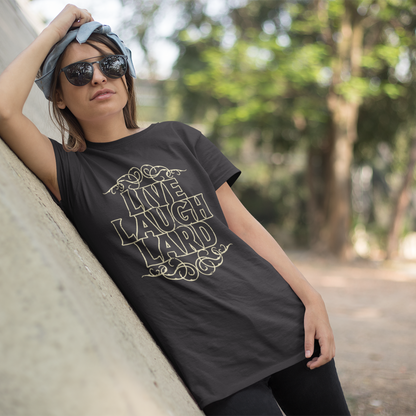 A woman wearing a black graphic t-shirt with 'LIVE LAUGH LAUGH' printed in gold, leaning against a concrete wall in an outdoor park setting.