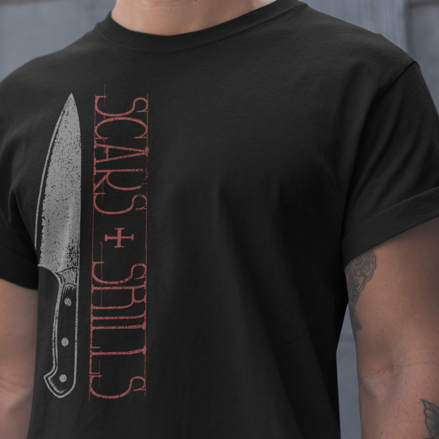 A man wearing a black t-shirt with a graphic print of a knife and the words 'SCARS + SHILLS' in distressed red and gray lettering.