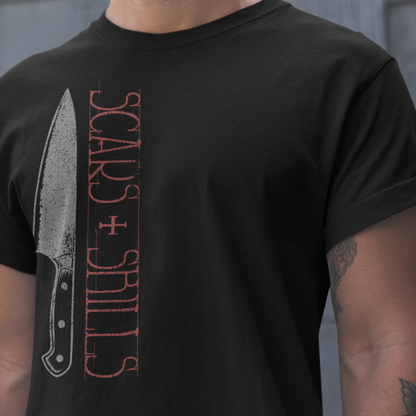 A man wearing a black t-shirt with a graphic print of a knife and the words 'SCARS + SHILLS' in distressed red and gray lettering.