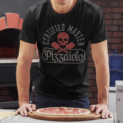 A man wearing a black graphic t-shirt with a red and white 'Certified Master Pizzaiolo' skull and cross-axes design, standing behind a pizza on a wooden board in front of a brick oven.