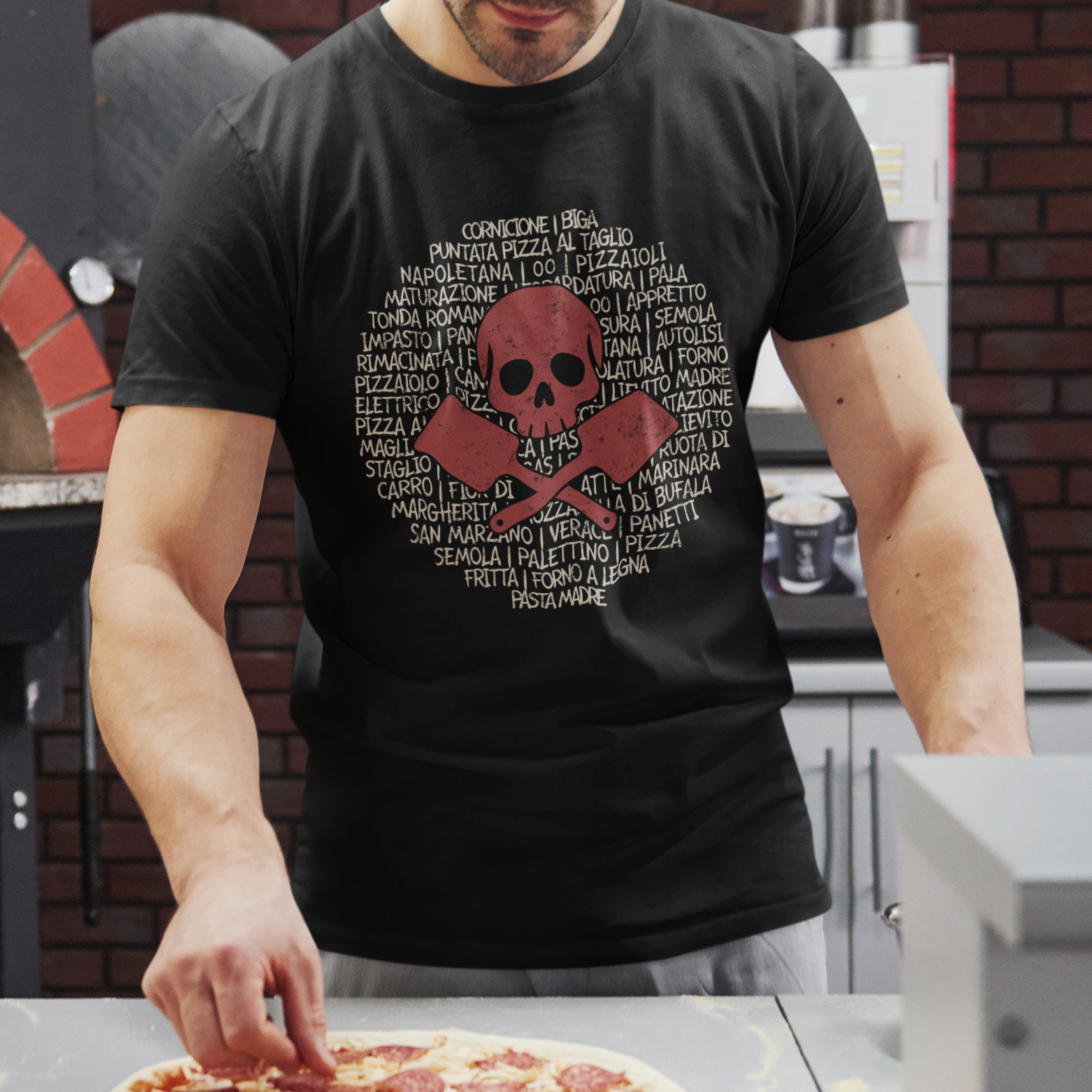 A man wearing a black graphic t-shirt with a red skull and crossbones design over a circular list of pizza toppings, standing in a kitchen and reaching for a pizza on a counter.
