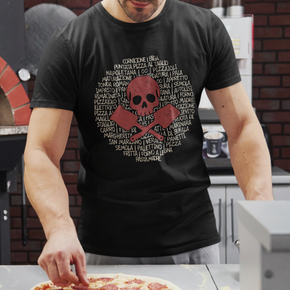 A man wearing a black graphic t-shirt with a red skull and crossbones design over a circular list of pizza toppings, standing in a kitchen and reaching for a pizza on a counter.