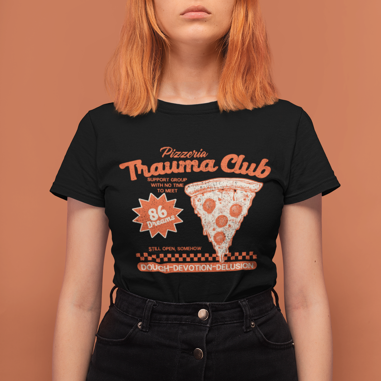 A model with orange hair wearing a black graphic t-shirt that reads 'Pizzeria Trauma Club' with a pizza slice illustration, posed against a solid terracotta background.