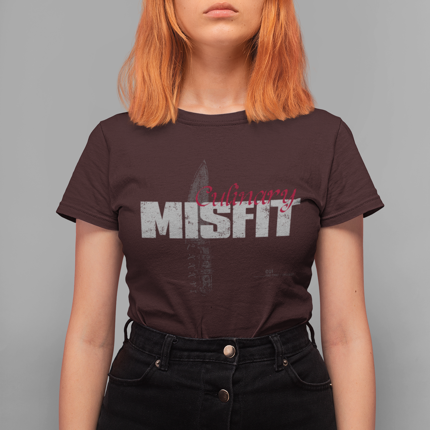 A model with bright orange hair wearing a dark brown Misfit band t-shirt and black high-waisted jeans, standing against a solid gray background.