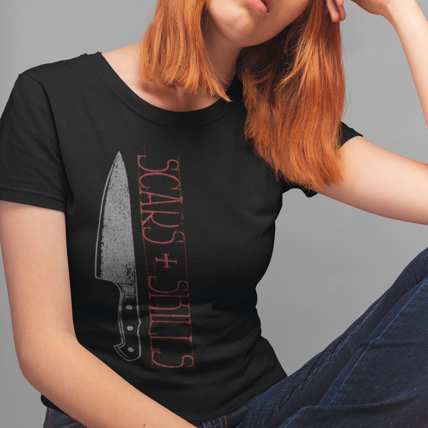A woman with strawberry-blonde hair wearing a black graphic t-shirt with a knife illustration and the text 'SCARS + STITCHES' in red, posed against a solid gray studio background.
