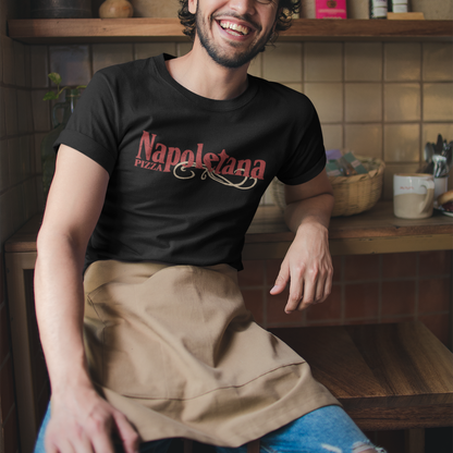 A smiling man wearing a black t-shirt with a red 'Napolesta 1927' graphic, a beige apron, and ripped blue jeans, sitting in a rustic kitchen setting.